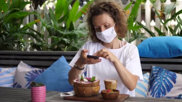얼굴에 마스크를 쓴 백인 여자가 건강하고 맛있는 스무디 그릇 사진을 찍고 있습니다. 세계적 유행병중의 불안 한 나날들 — 비디오