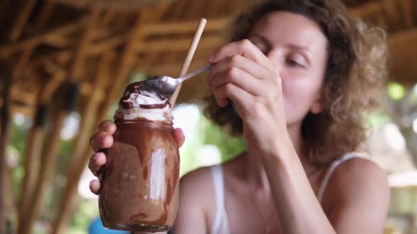 Chica rubia intenta crema batida de batido de chocolate y lo disfruta con los ojos cerrados. Postres en un café de playa — Vídeos de Stock