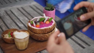 Smoothie hindistan cevizi kasesinin vejetaryen kahvaltısının en üst görüntüsü ahşap tahtada servis ediliyor ve sütsüz latte akıllı telefon ile fotoğraflanıyor. 