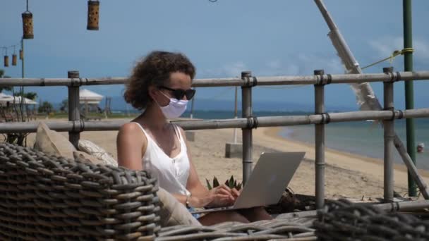 Távoli munka a parton a koronavírus alatt. Nő arcmaszkban a tengerparti étteremben — Stock videók