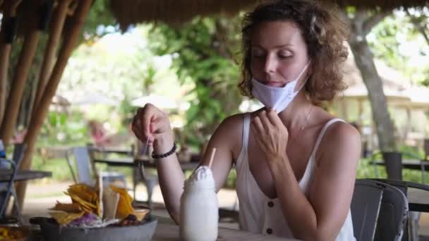Menina branca tira sua máscara protetora para tentar chantilly de seu milkshake durante o almoço em um restaurante ao ar livre. — Vídeo de Stock