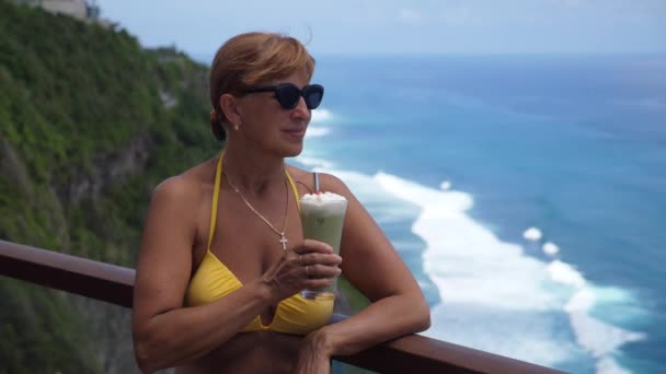 La meilleure vie à la retraite. S'adapter femme d'âge moyen dans un maillot de bain boissons cocktail crémeux profiter de la vue sur l'océan depuis le balcon falaise — Video