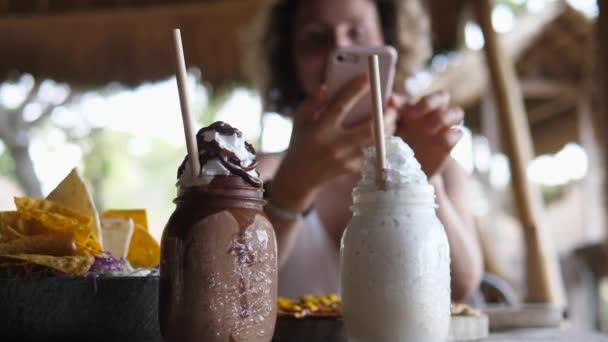 Ванильные и шоколадные молочные коктейли в стеклянных банках со взбитыми сливками и бумажными соломинками на переднем плане. Женщина фотографирует его на заднем плане — стоковое видео