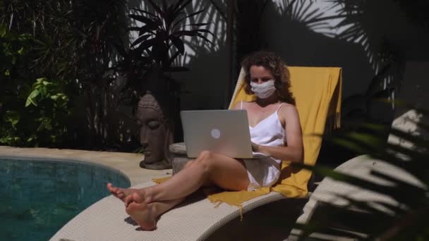 Молодая женщина в защитной маске работает на ноутбуке, загорая у бассейна. Концепция удаленных рабочих отпусков. — стоковое видео