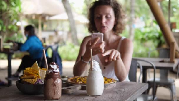Белая девушка в пляжном баре разговаривает и показывает на камеру еду на столе. Оставаться на связи с близкими — стоковое видео