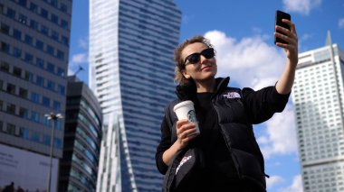 Genç bir kadın elinde Starbucks bardağıyla gökdelenlerin önünde selfie çekiyor. Varşova-Kasım-2020