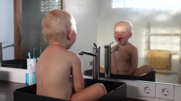 Bayi Kaukasia kecil yang lucu duduk di wastafel bermain dengan air melihat di cermin — Stok Video