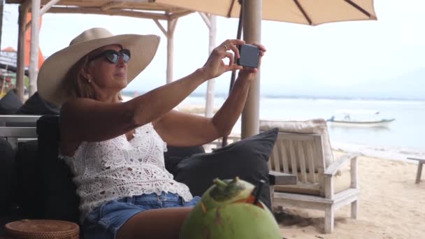 Donna di mezza età che fotografa l'oceano mentre si gode la vista dal divano del beach club — Video Stock