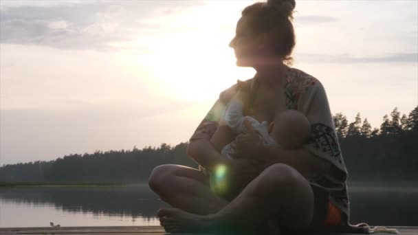 Jonge moeder borstvoeding haar baby op de kade van het meer bij zonsondergang. Moeder baby bonding tijd. — Stockvideo