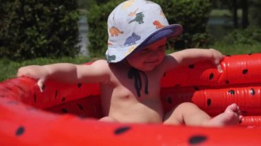 Cute baby in bucket hat relaxing in inflatable watermelon-style swimming pool. High quality FullHD footage