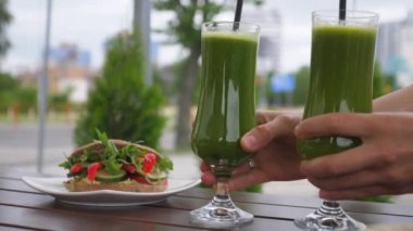 Hands serving two green smoothies with straws in long glasses. Vegan sandwich on the background. Healthy life style concept 