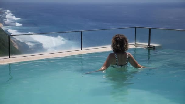 Vivere la vita migliore. Una donna salta fuori da una piscina a sfioro e spruzza acqua. Oceano e montagne sullo sfondo — Video Stock