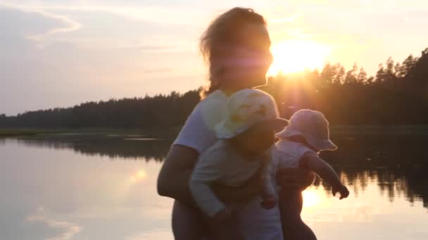 Feliz jovem mãe segurando e girando seus gêmeos bebê ao pôr do sol. Lago e árvores no fundo. Férias em família. — Vídeo de Stock