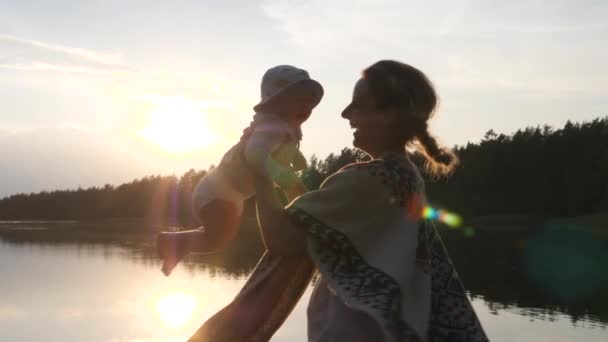 Boldog fiatal anya fogja és pörgeti a babáját vödör kalapban naplementekor. Tó és fák a háttérben. — Stock videók