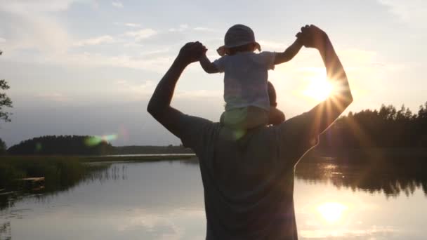 Boldog baba ül az apja vállán közvetlen napfényben. Tó és fák a háttérben. — Stock videók