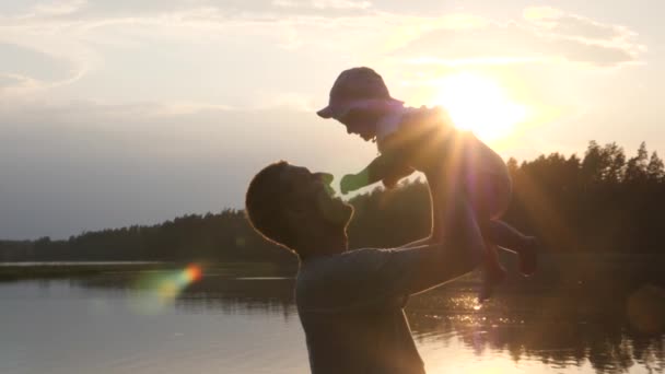 Happy young father lifting up his baby at sunset. Lake and trees on the background. Single parenting concept. — Stok video