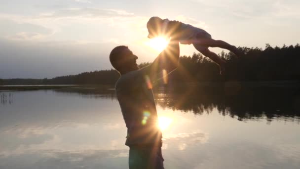 Šťastný mladý otec při západu slunce točí svým dítětem v kbelíkové čepici. Jezero a stromy na pozadí. Rodinná dovolená. — Stock video