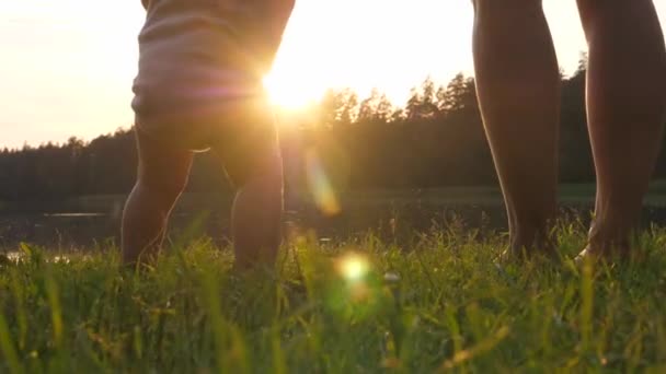 Orang tua dan bayi berdiri bertelanjang kaki di rumput saat matahari terbenam di tepi danau. Konsep induk tunggal. — Stok Video