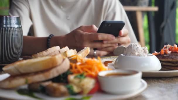 Hands browsing on the phone on the background in the focus. Blurred foreground with the breakfast — Stock Video