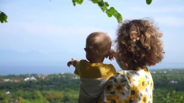 Bir kadının bebeğini kucağına alıp tepenin tepesinden ormanları göstermesi. Sarışın bebek çok uzakları işaret ediyor 