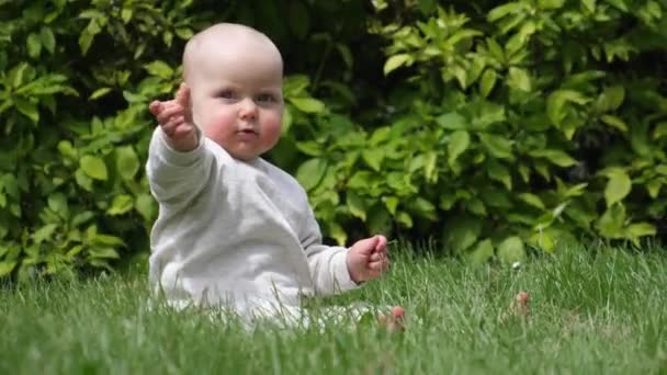 Parktaki sevimli bebek çimlerin üzerinde oturmuş kameraya el sallıyor. — Stok video