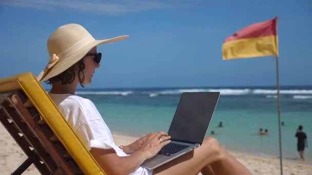 Oldalnézet egy nő szalmakalapban ül egy nyugágyon egy strand típusok a laptopján — Stock videók