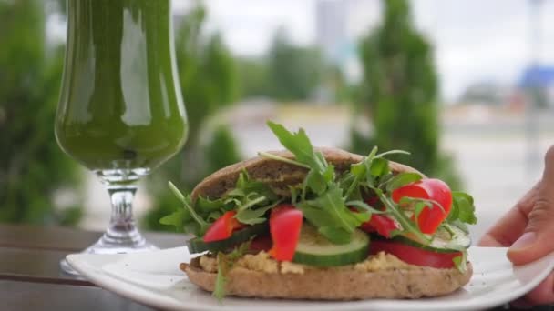 Mano que sirve sándwich vegano orgánico saludable en la mesa de madera marrón. Batido verde con paja n vaso largo en la parte posterior. Concepto de estilo de vida saludable — Vídeos de Stock