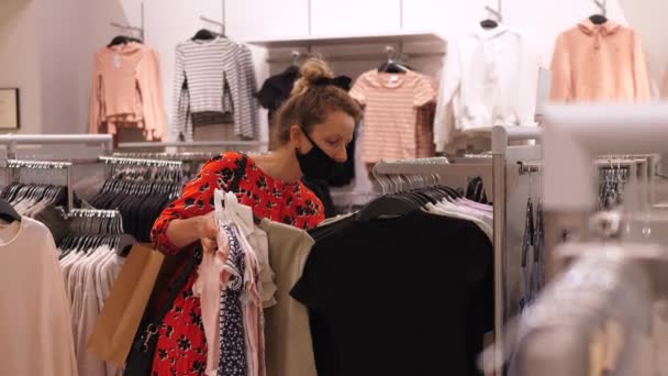 Flicka bär svart ansiktsmask shopping kläder efter karantän i köpcentret. Hangers på hyllan. Säker shopping under COVID-19-pandemin. — Stockvideo