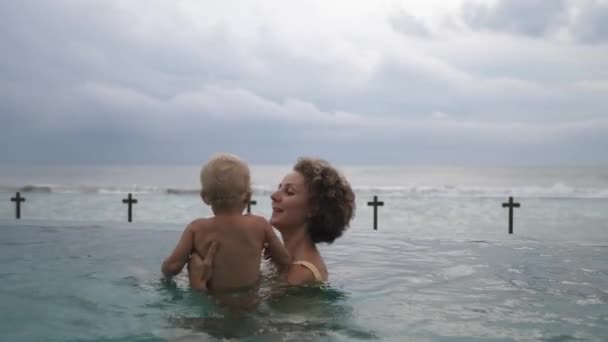 Jonge blanke moeder speelt met haar baby in het zwembad met uitzicht op een oceaan. Gelukkige jeugd concept — Stockvideo