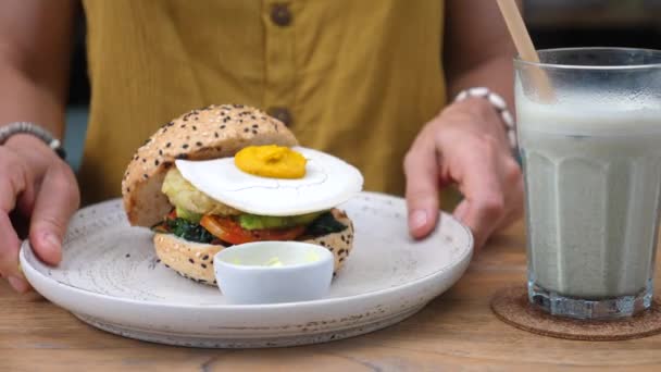 Oběd v americkém bistru ve veganském stylu. Samice přináší na stůl rostlinný burger s veganským vejcem. Mléčný koktejl s papírovou slámou ve sklenici — Stock video