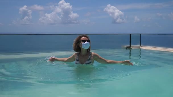 Viajar durante la pandemia de covid-19. Mujer caucásica con mascarilla en una piscina infinita — Vídeo de stock