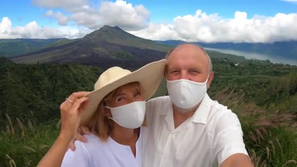 Middle-aged couple in face masks taking a selfie with the mountains and ocean in the background. Romantic getaway during covid-19 pandemic — Stock Video