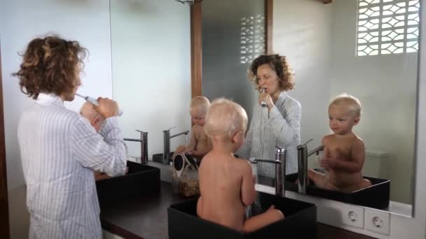 若い母親は子供の双子を流しに座って彼女の歯を磨く。楽しさと教育活動 — ストック動画
