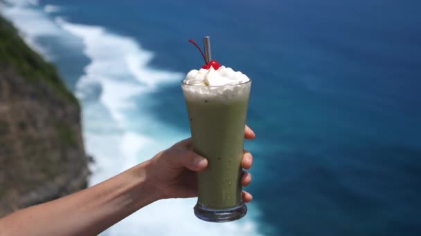 Hand holding iced matcha green tea with whipped cream and a cherry on top with the ocean line on a background. — Stock Video