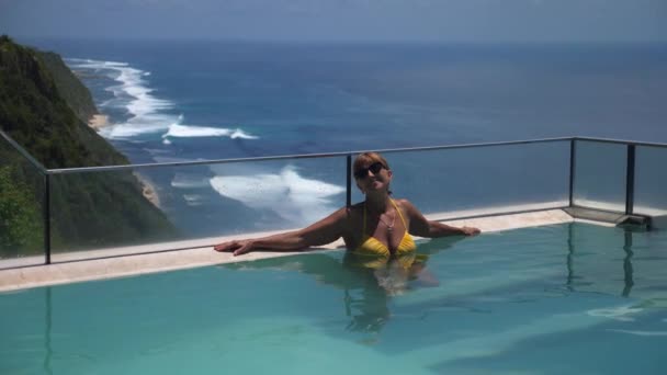 Mujer de mediana edad sonriendo a la cámara mientras se relaja en una piscina infinita con vistas a un océano — Vídeo de stock