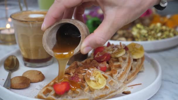 Desayuno vegano saludable y delicioso. Primer plano de la mano verter miel en gofres servidos con leche de café sin lácteos. — Vídeos de Stock