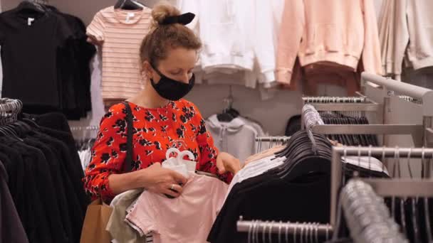 Дівчина з булочкою для волосся в чорній масці для обличчя, що проходить через одяг у модному магазині після закінчення карантину. Безпечні покупки під час пандемії COVID-19 . — стокове відео