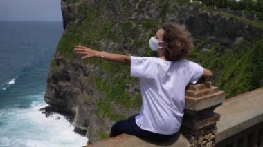 Corona virüsü sırasında tek kişilik yolculuk. Kafkasyalı bir kızın yüz maskesi taktığı bir uçurumun kenarında oturup kollarını açtığı görüntüler..