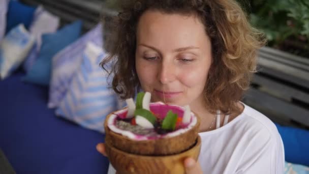 Chica rizada mira de cerca con emoción en el tazón de batido tropical decorado con bayas y chips de coco tostados servidos en cáscara de coco — Vídeo de stock