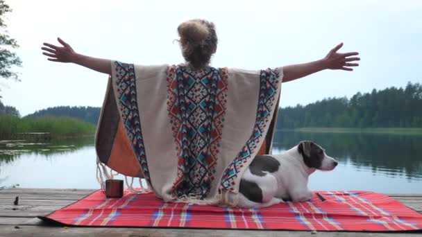 Жінка в затишному пончо широко розтягує руки, цінуючи природу і час з білим і коричневим собакою на причалі озера на заході сонця. Концепція самодопомоги . — стокове відео