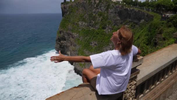 Mujer de mediana edad sentada en un banco de un acantilado extiende los brazos apreciando ampliamente la vista al mar. Vida después de la jubilación — Vídeos de Stock