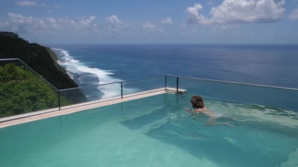 Concepto de resorts de lujo. Mujer joven nadando en una piscina infinita con la impresionante vista de un océano — Vídeo de stock