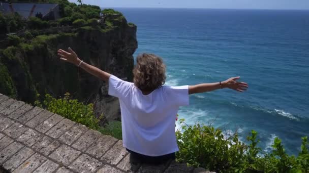 Pohled zezadu na ženu spojující se s přírodou, oceánem a horami sedící na vrcholku útesu a bojující s rukama — Stock video