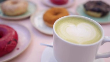 Üzerinde kalp tasarımı olan beyaz porselen bir fincan vejetaryen matcha latte getir. Bulanık arkaplanda farklı donut türleri