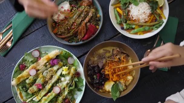 Top kilátás az emberek eszik az egészséges vegán ételeket evőpálcikával. Tálak tele színes zöldségekkel, fekete, vaskos fa asztalon tálalva. — Stock videók