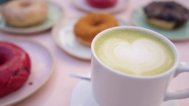 Κοντινό πλάνο του λευκού φλιτζάνι πορσελάνη vegan matcha latte με το σχεδιασμό της καρδιάς. Διαφορετικοί τύποι ντόνατς στο θολό φόντο — Αρχείο Βίντεο