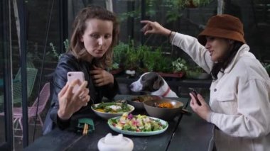 İki kız hippi kafeteryasında yemek yerken sevimli kahverengi ve beyaz bir köpekle selfie çekmeye çalışıyor.