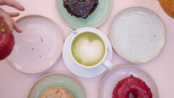 Ciambelle vegane assortite servite intorno a una tazza di caffellatte matcha sul tavolo rosa pastello. Due mani che prendono le ciambelle dai piatti e le rimettono a morso — Video Stock