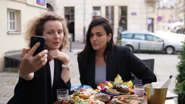 Twee jonge vrolijke zakenvrouwen nemen selfie met een smartphone tijdens hun lunch buiten — Stockvideo