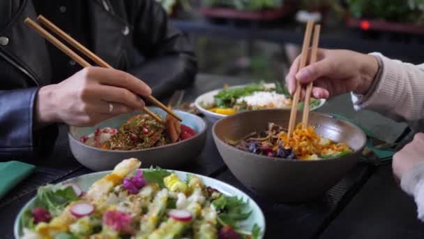 Dos personas con palillos disfrutando de sus cuencos llenos de comidas saludables al estilo de la cocina asiática Cena servida en una mesa de madera negra y gruesa — Vídeos de Stock